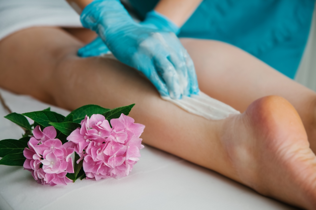 Zwei nackte Beine, zwei rosarote Blüten und eine Hand mit blauen Hygienehandschuhen die ein weißes Tuch auf ein Bein hält