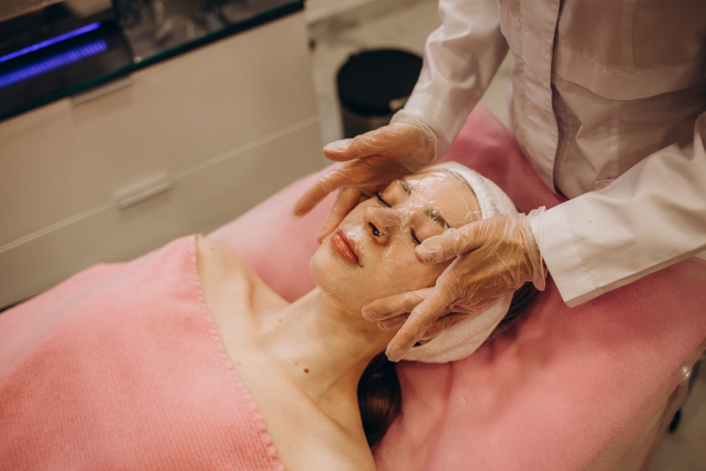 Frau liegt auf einer Kosmetikliege und hat eine Maske auf ihrem Gesicht die von zwei Händen mit Hygienehandschuhen glatt gestrichen wird